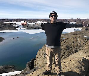 Expeditioner stands on a peak high above a vast blue lake surrounded by brown rocky hills