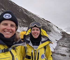 Dressed in yellow outer layer clothing, two female scientists stop for a photo below a sandy, snowy hill