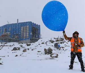 An expeditioner holds a blue weather balloon in front of the blue Met building while snow swirls around.