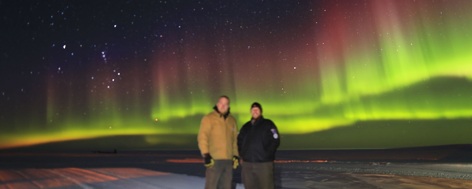 Two expeditioners stand on the ice in front of a colourful Aurora Australis in the background