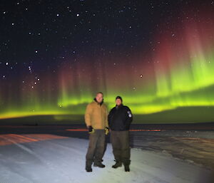 Two expeditioners stand on the ice in front of a colourful Aurora Australis in the background