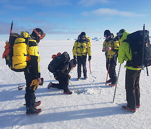 An expeditioner use a measuring tape to record the sea ice depth while the rest of the group watches on enthusiastically
