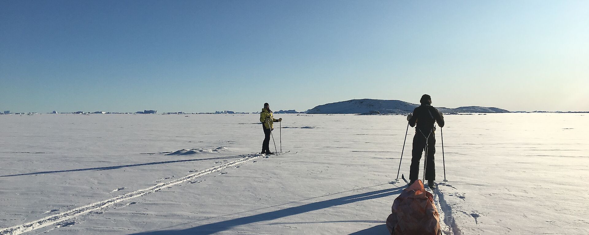 Two expeditioners ski towards Anchorage Island across snow covered sea ice into the low sun and blue sky