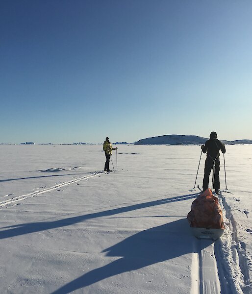 Two expeditioners ski towards Anchorage Island across snow covered sea ice into the low sun and blue sky