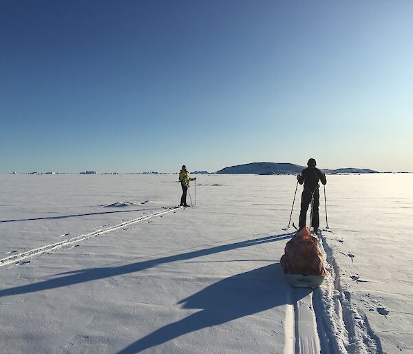 Two expeditioners ski towards Anchorage Island across snow covered sea ice into the low sun and blue sky