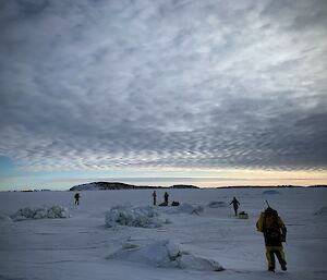 A group of five navigates the initial entry point from land onto the grey sea ice under a cloudy sky