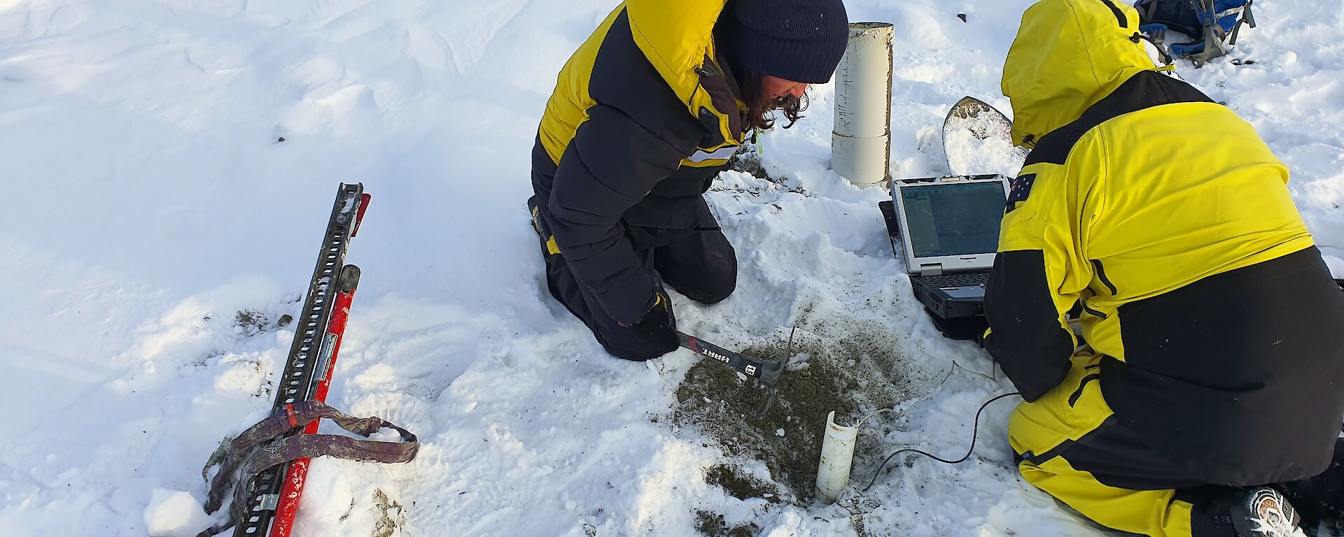 Two expeditioners sit on the snowy ground while taking measurements from a soil probe