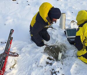 Two expeditioners sit on the snowy ground while taking measurements from a soil probe