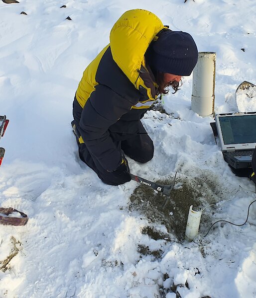 Two expeditioners sit on the snowy ground while taking measurements from a soil probe