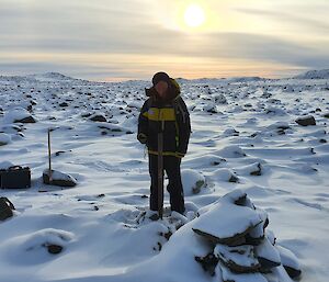 An expeditioner at a site surrounded by snowy rocky landscape and low sun in background