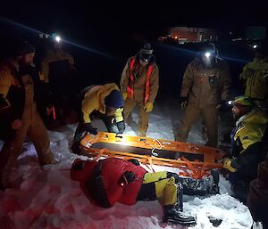 Doctor prepares to lift the casualty into an orange stretcher, in the dark surrounded by rescuers