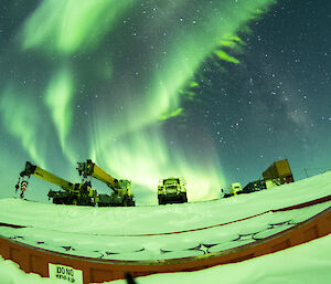 A huge green aurora bursts through the sky above the parked cranes and trucks
