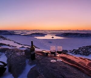 A bottle of wine and two glasses in front of a colourful sunset over the ice.