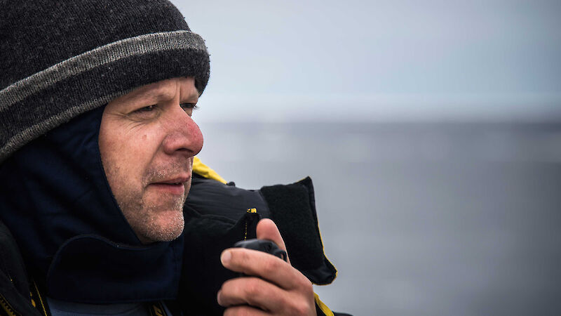 A man wearing a beanie looks out observing the ocean holding a radio to his mouth