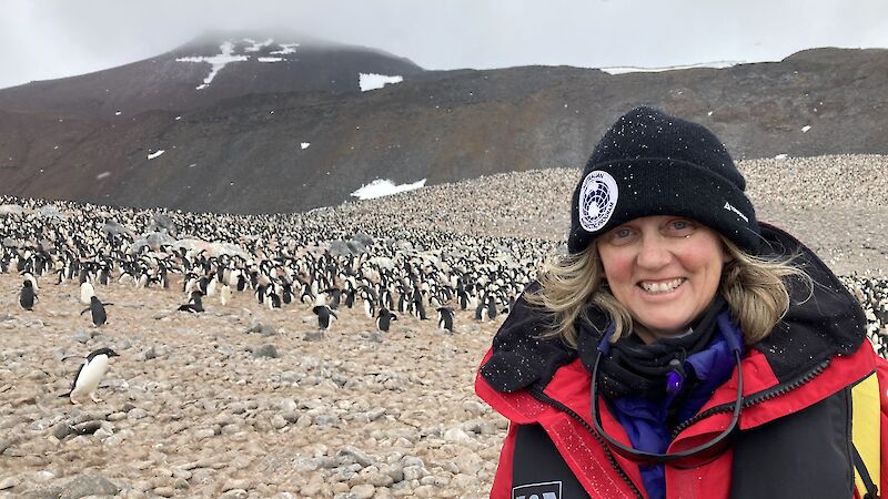 A woman in a red jacket and black beanie stands in front of a penguin colony