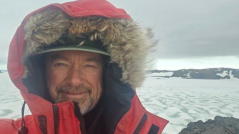 Head and shoulders of a man wearing a hooded red jacket with sea ice and rocky outcrops in the background