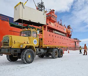 A Mack truck on the sea ice beside the icebreaker Aurora Australis, with a crane lowering a shipping container onto its flat deck.