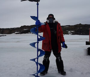 A scientist with a large post hole drill, beside a hole in the sea ice.
