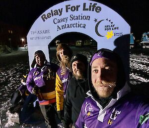 Four expeditioners stand in front of an arch at night