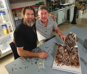 Two men sitting next to a birthday cake
