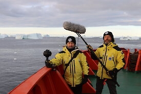 John McCormick holds a 360 degree camera and Adam Nash holds a boom microphone, on the deck of the Aurora Australis in Antarctica.