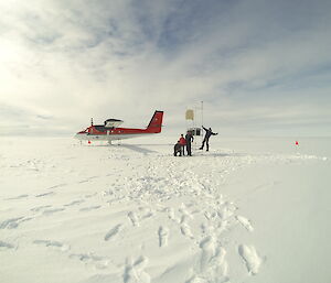 Three expeditioners and a red plane on a flat, ice covered landscape