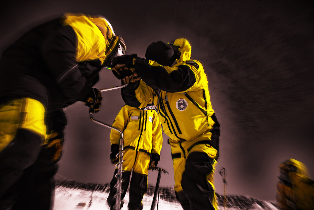 Three people wearing yellow jackets holding a piece of equipment with a dark sky behind them.