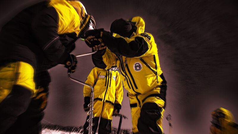 Three people wearing yellow jackets holding a piece of equipment with a dark sky behind them.