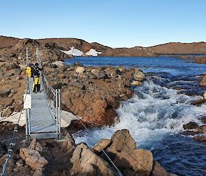 an expeditioner gazes out over watts hut bridge during the bright summer as strong rapids flow underneath