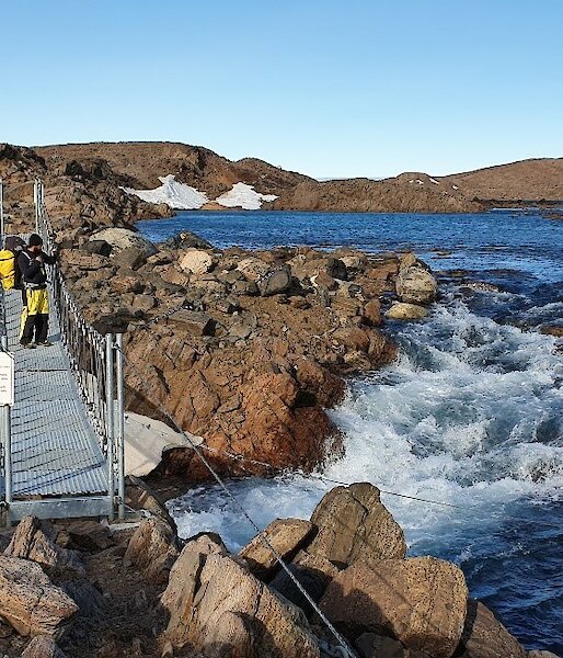 an expeditioner gazes out over watts hut bridge during the bright summer as strong rapids flow underneath