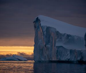 A large iceberg dominates a moody frame of orange-grey sky and blue-grey water