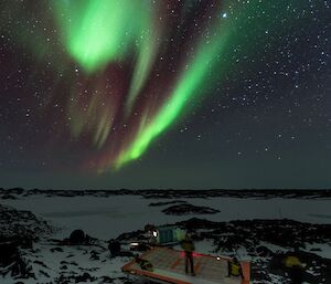 An aurora lights up the sky above a field hut while the group watch on from the helipad