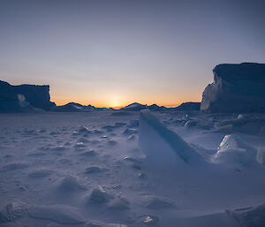 The hidden orange sun lights up a valley of icebergs and blue, crumbled ice chunks