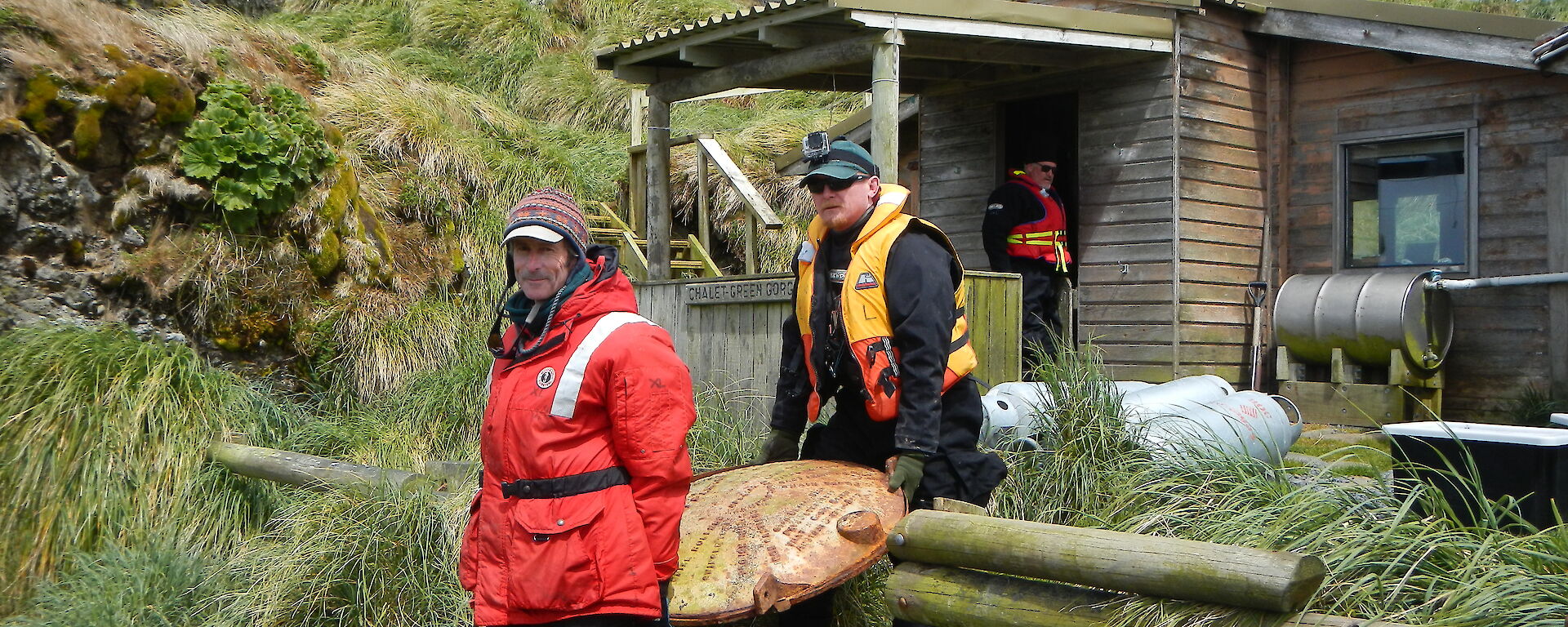 Two expeditioners carry a water tank end out from a field hut