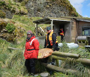 Two expeditioners carry a water tank end out from a field hut