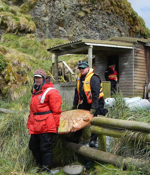 Two expeditioners carry a water tank end out from a field hut