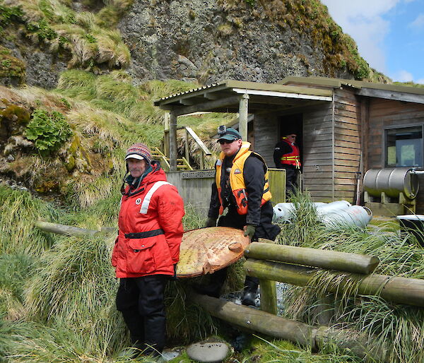 Two expeditioners carry a water tank end out from a field hut