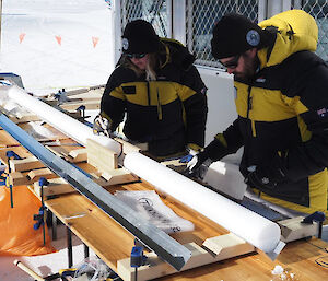 Two people marking ice core sections with pencils. The ice cores are sitting in a v-shaped channel on a wooden bench.