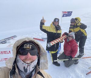 4 smiling expeditioners stand on the ice holding an ice core. An Australian flag flies in the background.