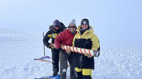 3 expeditioners hold a long tube in an icy landscape.