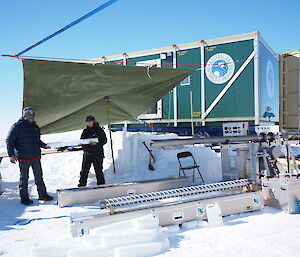 2 expeditioners carry cylinders of ice amongst a field camp set up on the ice.