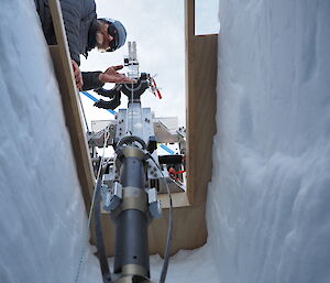 An expeditioner lowers a drill into a trench cut out of the snow.