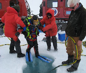 Three people helping two divers into an ice hole.