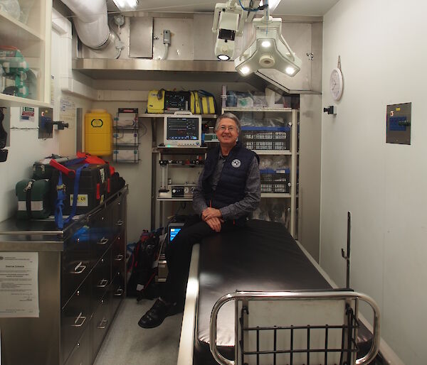 Dr Strauss sitting on the edge of the operating table with medical equipment behind him and operating theatre lights above.