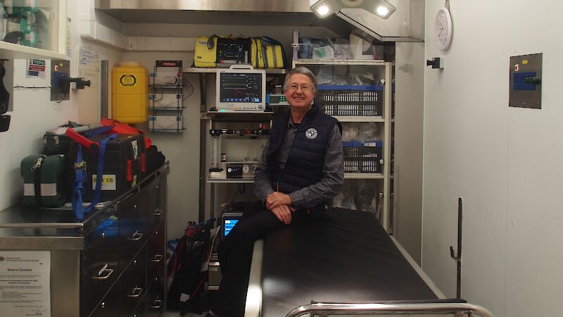 Dr Strauss sitting on the edge of the operating table with medical equipment behind him and operating theatre lights above.