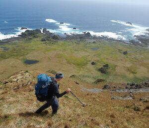 A woman with a large pack on her back walks along the slope edge above the ocean