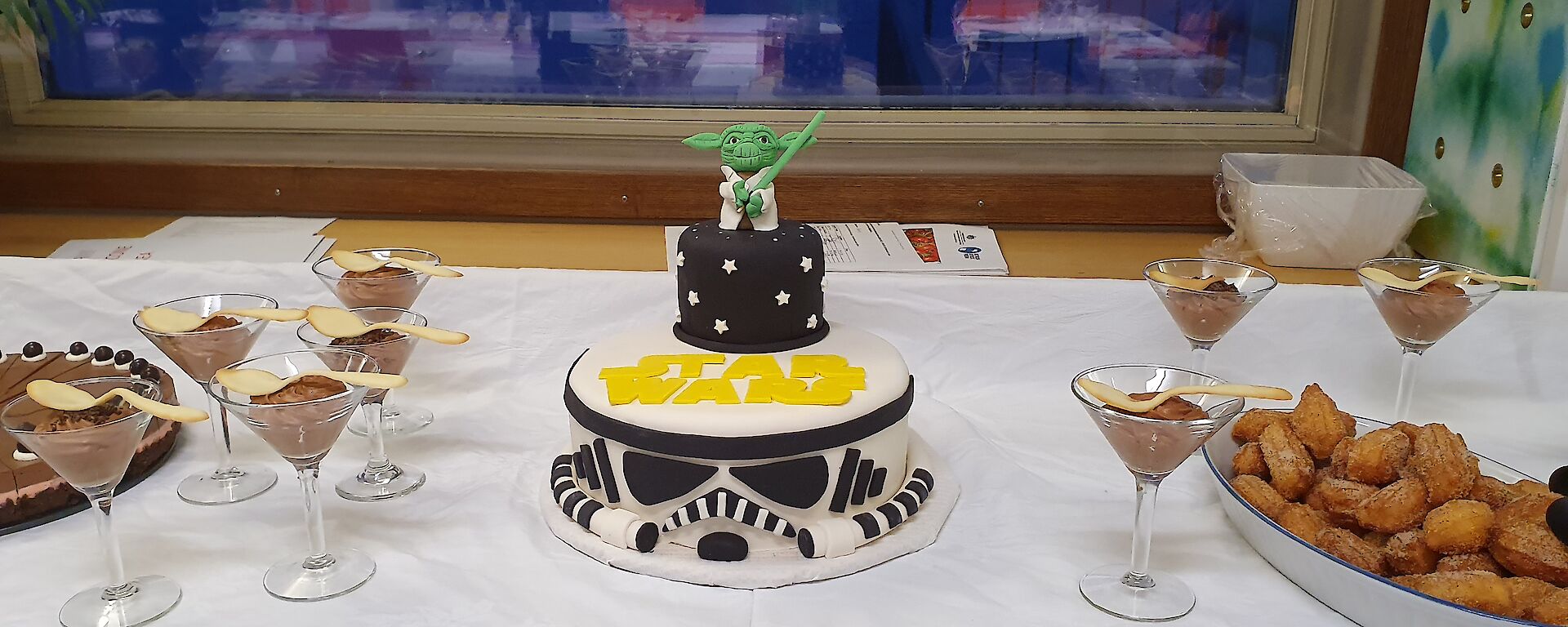 A Starwars themed cake is flanked on the dessert table by churros on one side and chocolate mousse on the other