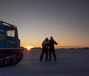 Two expeditioners standing on the ice next to a Hagg, enjoying the sunrise