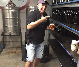A man holding a pint of beer grins and points to more bottles on the shelf