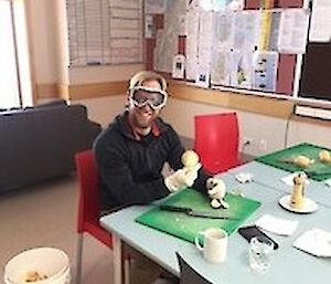 A man sits at a table peeling onions whilst wearing protective goggles